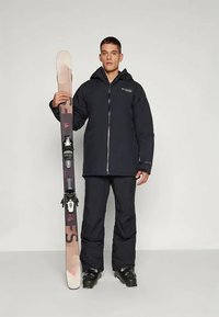 Black insulated jacket with a front zipper and logo, paired with matching black ski pants. Individual holds pink and brown skis with bindings.
