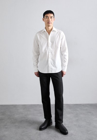 Vit långärmad skjorta med knäppning och lätt rutmönster, kombinerad med mörka slim-fit byxor och svarta ankelskor. Enkel, ren design.
