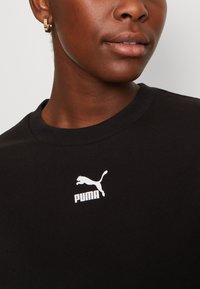 Černé bavlněné tričko s kulatým výstřihem; na levé straně hrudi má bílé logo Puma potištěné sítotiskem. Hladká textura.