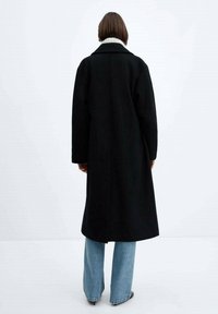 Lungo cappotto nero con una texture simile alla lana e un ampio colletto, caratterizzato da un dettaglio interno a collo di maglia bianca. Indossato sopra jeans azzurri chiari.