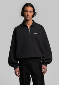 Sweatshirt noir à col zippé quart de tour, avec un logo blanc sur la poitrine et une coupe décontractée. Fabriqué en tissu texturé.