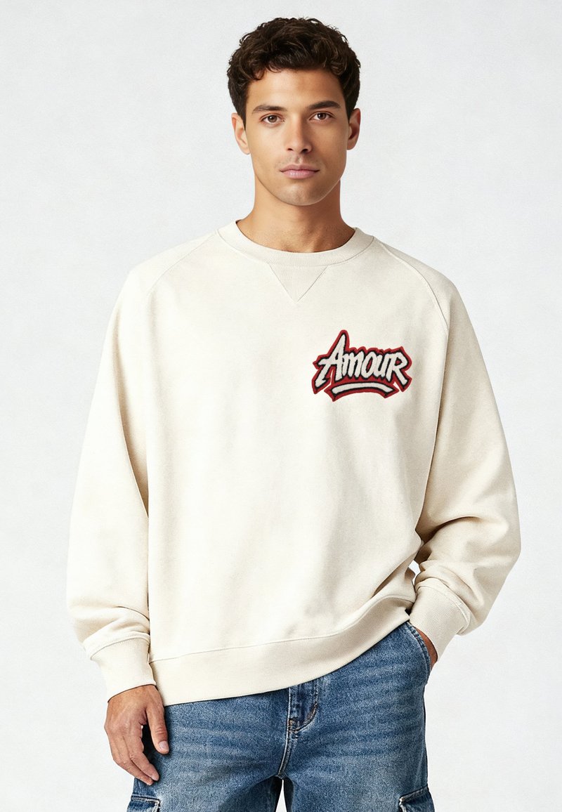 Sweat-shirt beige clair à manches raglan, avec broderie "Amour" en rouge et noir sur la poitrine. Texture douce et coupe décontractée.