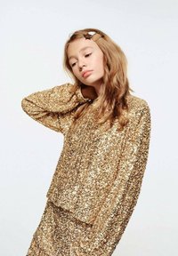 Top con paillettes dorate, maniche lunghe, vestibilità ampia e superficie testurizzata. Decorato con una molletta per capelli. Sfondo semplice.