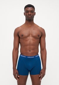Blaue Baumwoll-Boxershorts mit enger Passform, ausgestattet mit einem weißen Logo-Bund und einer glatten Textur. Das Modell ist oberkörperfrei.