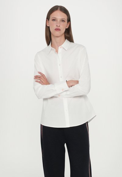 Camicia bianca con bottoni realizzata in tessuto morbido, caratterizzata da un colletto classico, maniche lunghe e un orlo leggermente curvato.