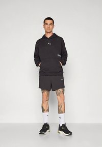 Camisola preta com capuz, bolso canguru e logótipo, combinada com calções escuros. Modelo usa sapatos desportivos pretos e meias brancas. Textura do tecido lisa.