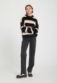 Femme debout, bras croisés, portant un pull duveteux rayé noir, blanc et rose, un jean noir et des chaussures noires et blanches.