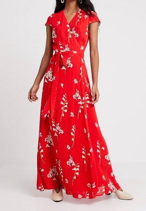 Vrouw die een rode maxi-jurk met bloemenprint draagt, met korte mouwen, een tailleband en beige puntige schoenen.