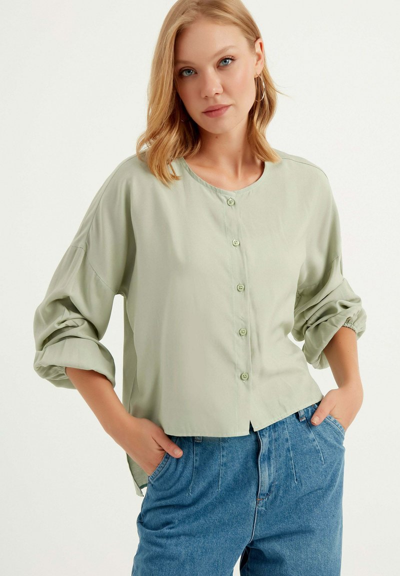 Blusa verde chiaro con bottoni e scollo rotondo, maniche arrotolate e vestibilità rilassata, abbinata a pantaloni in denim blu a vita alta.