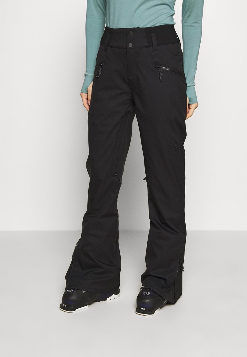 Personne portant un pantalon de neige noir zippé et des bottes de ski noires, debout devant un fond clair uni.