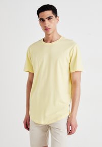 Šviesiai geltona trumpomis rankovėmis marškinėliai su apvalia iškirpte, pagaminti iš minkštos medvilnės, pasižymintys laisvesne forma ir išlinkusiu apačios kraštu.