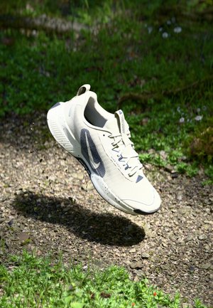 Lys beige atletisk sko med en tekstureret overdel, blå accenter, en polstret sål og et strømlinet design, hængende over en grusoverflade.