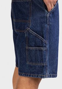 Ręka w kieszeni niebieskich dżinsowych szortów cargo z wieloma przeszywanymi kieszeniami, widocznych na udzie osoby na jednolitym jasnym tle.