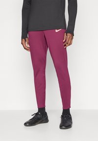 Calças de treino em cor bordô, feitas de tecido entrelaçado com um design afunilado. Apresenta um pequeno logótipo da Nike em dourado na parte da frente e calçado preto.