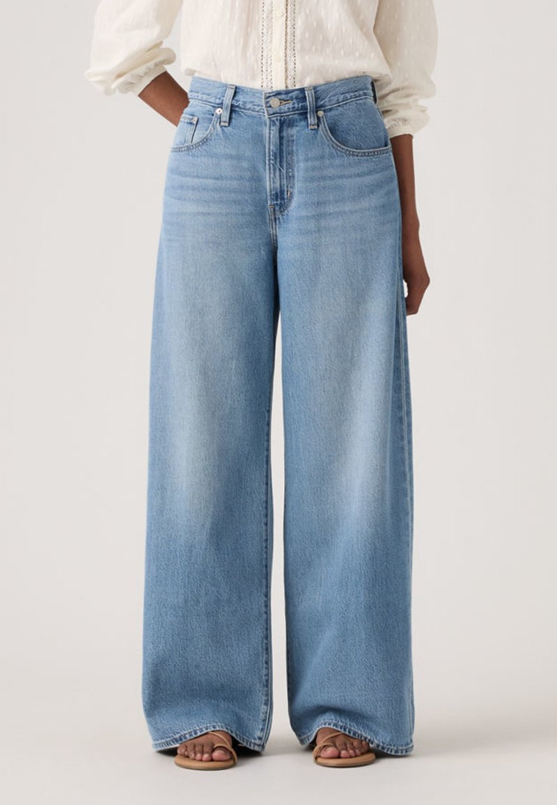 Person wearing light blue wide-leg jeans paired with a white long-sleeve blouse and brown sandals, standing with hands in pockets.