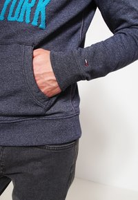 Sudadera gris oscuro hecha de un tejido texturizado. Presenta letras en azul brillante que dicen "NEW YORK" y un pequeño detalle en rojo, blanco y azul en la manga.