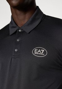 Polo noir en tissu texturé, avec une patte de boutonnage à trois boutons et un logo EA7 blanc sur la poitrine gauche. Design de col classique.