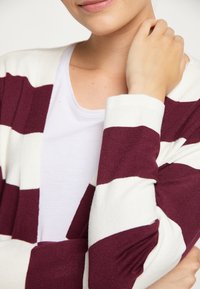 Maroon and cream striped cardigan in soft knit fabric, featuring a relaxed fit and open front, worn over a plain white shirt.