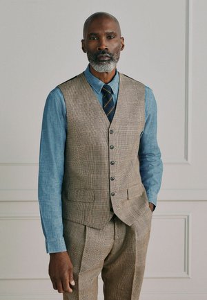 Man with gray beard wearing plaid beige vest and pants, blue denim shirt, and striped tie, standing with one hand in pocket.