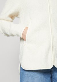 Bílá fleece bunda s texturovaným povrchem, se zapínáním na zip, bočními kapsami a měkkým interiérem, kombinovaná s modrými džínami.