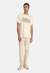 Lys cremefarvet t-shirt med "Petrol & Co" grafik, parret med lyse beige sweatpants og beige sneakers, der viser en afslappet pasform og blød tekstur.