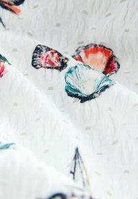 Biely textil so vzorom farebných mušlí v odtieňoch červenej, zelenej a modrej, s textúrovaným povrchom a malými bodkovanými detailmi.