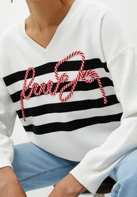 Pull en maille blanche à rayures horizontales noires, avec lettrage rouge style corde sur la poitrine. Col en V et poignets côtelés.