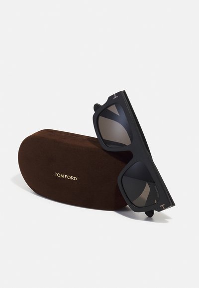 Lunettes de soleil noires rectangulaires avec verres dégradés, dotées d'une finition mate et d'un logo doré, exposées à côté d'un étui en daim marron.