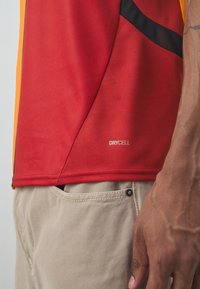 Camisa deportiva roja de poliéster con textura de malla, acentos en negro y naranja, y una etiqueta "DRYCELL" cerca del dobladillo. Combinada con pantalones color beige.