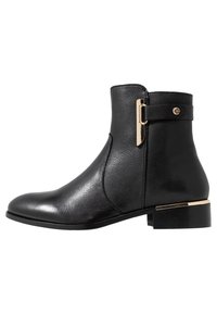 Bottine en cuir noir à texture lisse, bout arrondi et accent doré sur le talon. Bride latérale avec détail de boucle métallique.