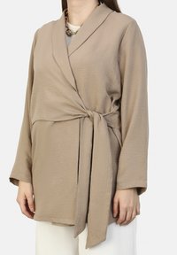 Beige wrap top with long sleeves and a tie closure. Textured fabric; features a shawl collar. Paired with light-colored bottoms.