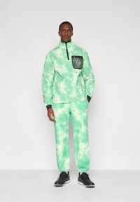 Πράσινο πουλόβερ με σχέδιο tie-dye, με μαύρη τσέπη με φερμουάρ και λογότυπο, σε συνδυασμό με παντελόνι με το ίδιο πράσινο σχέδιο tie-dye και μαύρα αθλητικά παπούτσια.