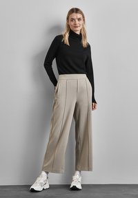 Beige vida byxor med veck, svart åtsittande polokrage och vita sneakers. Slät tyg och ren design mot en grå bakgrund.