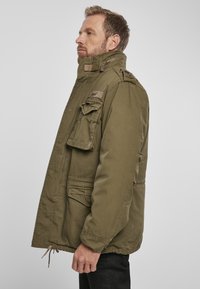 Olive green military-style jacket featuring a high collar, multiple pockets, a zip closure, and elastic cuffs. Textured fabric with visible stitching.