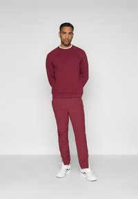 Felpa bordeaux con logo testurizzato e pantaloni taper coordinati. Sneakers bianche accentuano l'outfit con un contrasto. Tessuto morbido, vestibilità atletica.