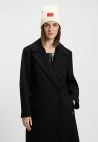 Cappotto di lana nera con lapel oversize e tasche laterali, abbinato a un berretto a coste crema con una toppa rossa.