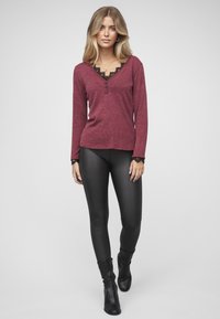 Bordeauxrotes Langarmshirt mit Spitzenbesatz und drei Knöpfen am Ausschnitt, kombiniert mit schwarzen Leggings aus Kunstleder und Stiefeletten.