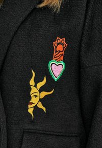 Čierna tkanina s farebnými vyšívanými motívmi: žltá tvár slnka a oranžová tigrova labka nad ružovým srdcom so zeleným obrysom.