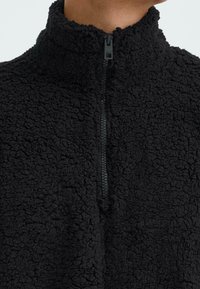 Sudadera de forro polar negra con cuello alto y media cremallera. Tejido texturizado con un aspecto suave y esponjoso, sin patrones ni acentos visibles.