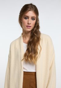 Kreminė spalvos kardiganas su minkšta, tekstūrine medžiaga ir atviru dizainu. Dėvima ant baltos palaidinės, derinama su rudos zomšos sijonu.