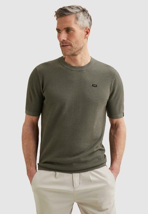 Mann mit kurzen grauen Haaren, der einen olivgrünen, strukturierten Kurzarmpullover und hellbeige Hosen trägt, mit den Händen in den Taschen.