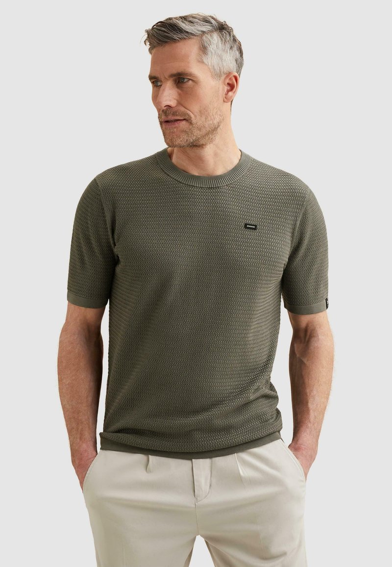 Homme aux cheveux gris courts portant un pull à manches courtes texturé vert olive et un pantalon beige clair, les mains dans les poches.
