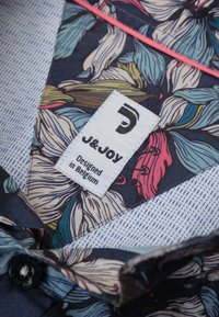 Stof met bloemmotief met donkerblauwe en kleurrijke bladeren. Inclusief een wit label met "J&Joy" en "Ontworpen in België." Knopdetail zichtbaar.