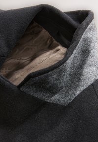 Gros plan sur le col d'un manteau en laine noire avec un accent en feutre gris et une doublure intérieure marron lisse et à motifs.