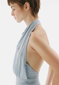 Vestido de cuello halter azul claro con suaves drapeados, espalda descubierta y cintura ajustada. La modelo luce unos grandes pendientes de oro en forma de aro. Textura de tejido suave.