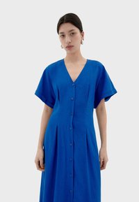 Vestito in lino blu con scollatura a V, maniche corte e chiusura con bottoni sul davanti. Presenta pieghe verticali per un tocco di dettaglio in più.