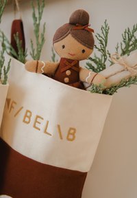 Vánoční punčocha z látky v krémové a tmavě červené barvě, obsahující plyšovou postavičku s hnědými vlasy a šaty, obklopenou zelení.