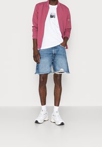 Rosa bomberjacka över en vit t-shirt, bärs med slitna denimshorts och vita sneakers med grå detaljer.