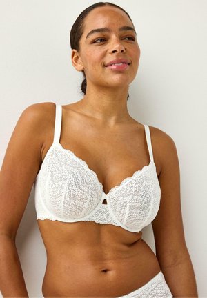 Nainen, jolla on tummat hiukset taaksepäin kammattuna ja valkoinen pitsirintaliivit päällä, seisoo yksinkertaisen vaalean taustan edessä, katsoo hieman sivulle ja hymyilee.