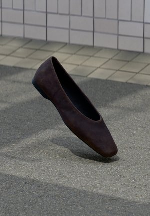 Viens tumši brūns plakanās kurpes pārvietojas virs teksturētas zemes ar flīzētu sienas fonu.